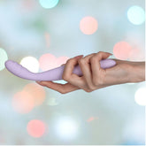 A hand holds the Svakom Cici 2 G-Spot Vibrator in Pastel Lilac, a light purple curved object made from liquid silicone, against a blurred background with pastel circular lights. The smooth texture and small ridges on one end indicate it's designed for precise G-spot pleasure.